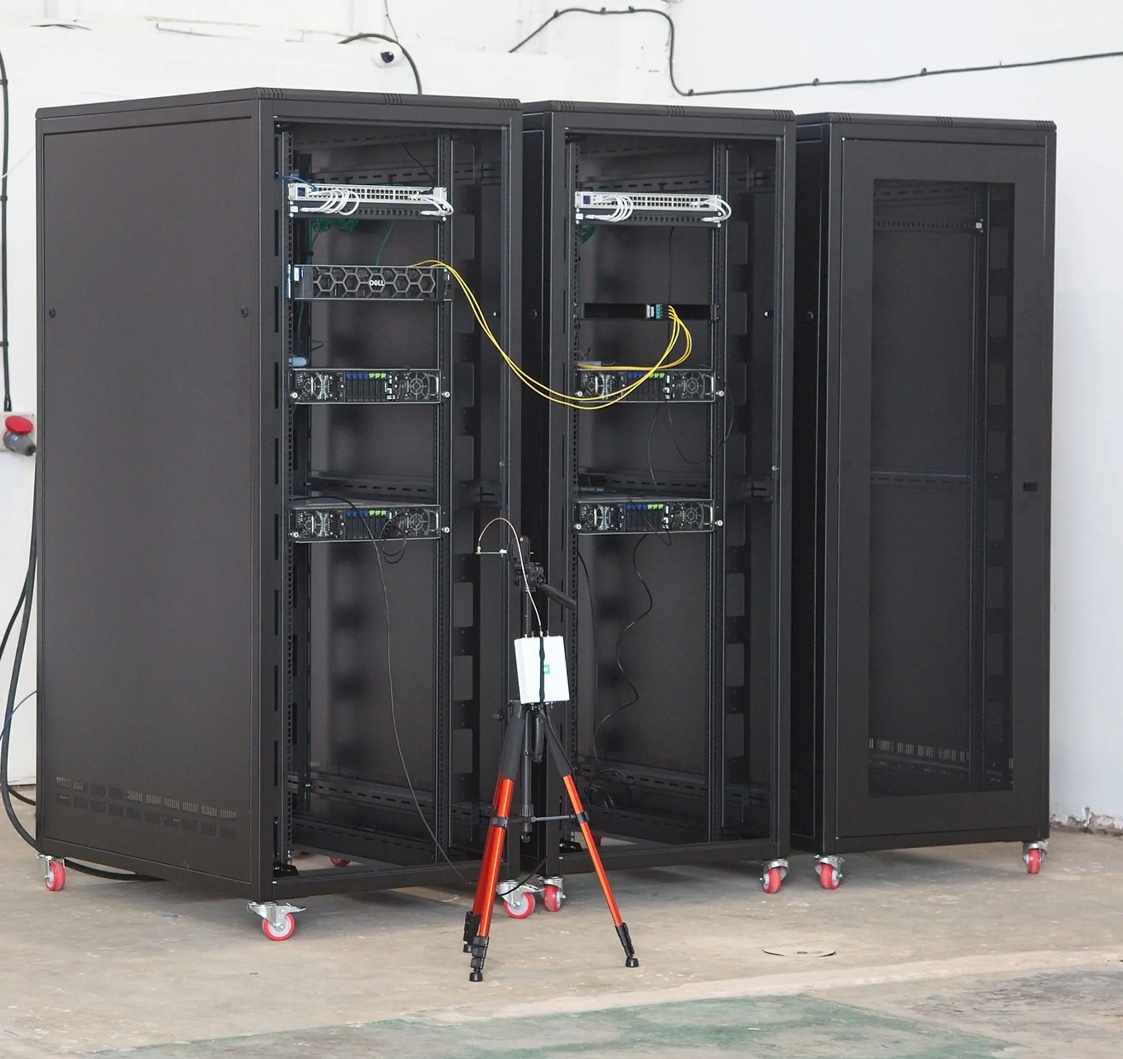 Photo of three open black server racks on castors in a workshop. The left two racks hold switches and rack-mount servers, with yellow fibre patch cables running between the racks. A camera on an orange tripod stands in front.
